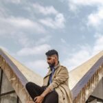 Fashionable young man with a full beard sitting on a modern urban structure outdoors.